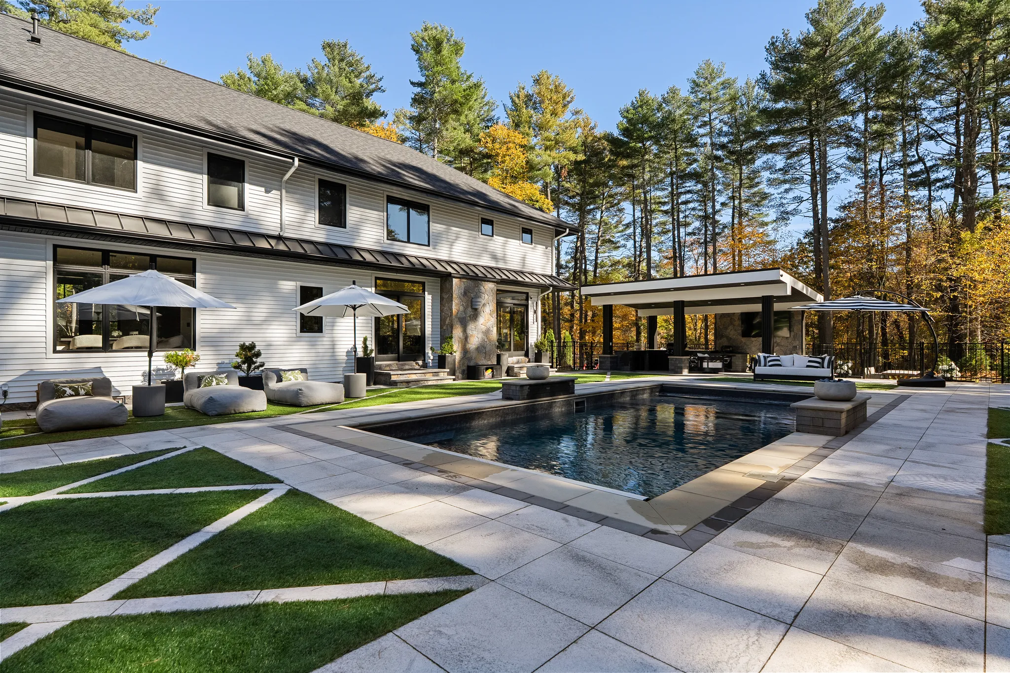 A modern backyard features a swimming pool, a large patio with outdoor seating, and a covered outdoor kitchen next to a house.