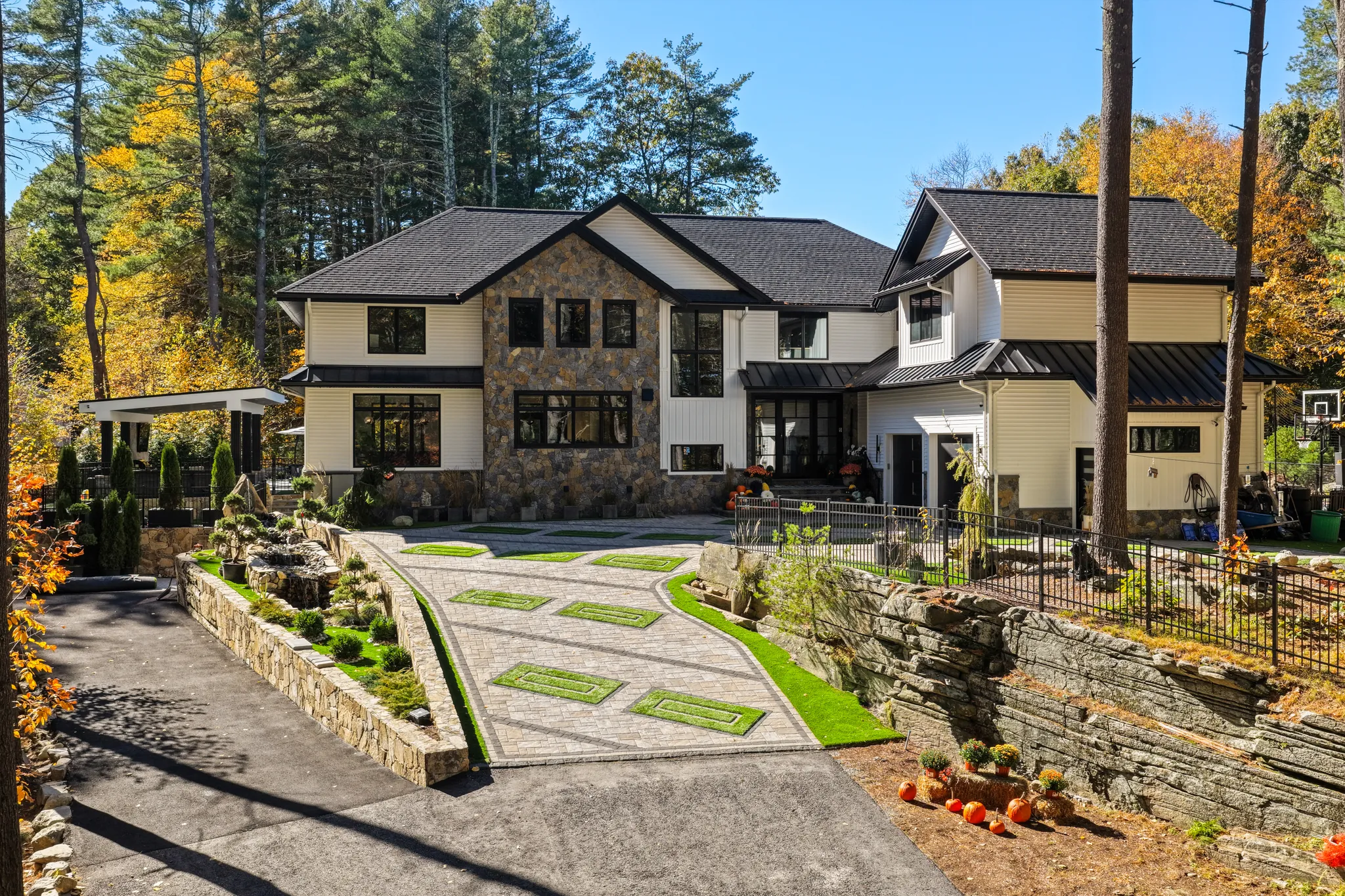 A modern house with stone and white siding features a paved driveway, extensive landscaping, and is surrounded by trees with autumn leaves.