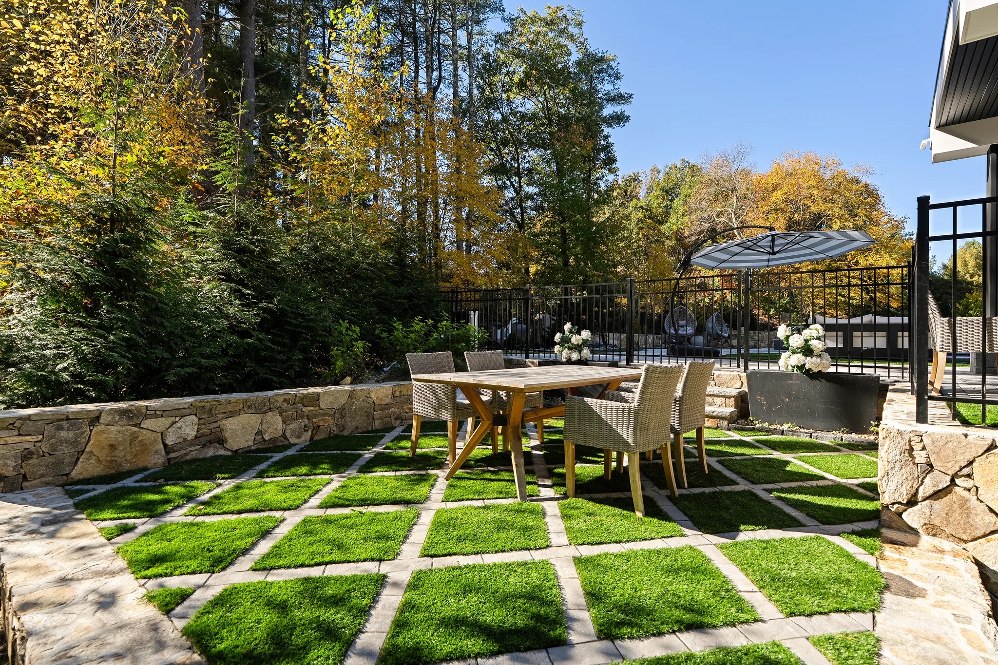 An outdoor dining area features a wooden table, wicker chairs, a stone wall, and grass pavers, bordered by a black fence and autumn trees.