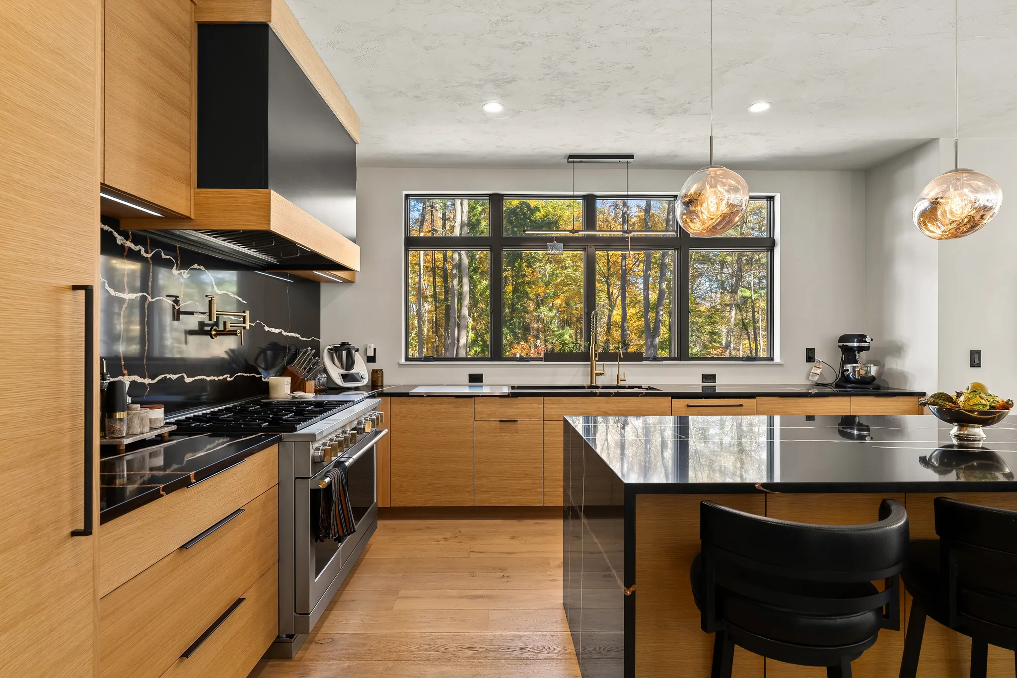 A modern kitchen features light wood cabinetry, black countertops, a large island, stainless steel appliances, and a window offering a view of a forest.