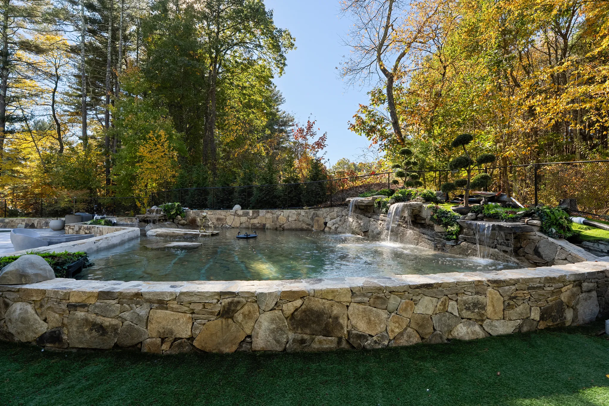 A naturalistic pond with waterfalls and a stone retaining wall is set in a lush yard with autumn trees.