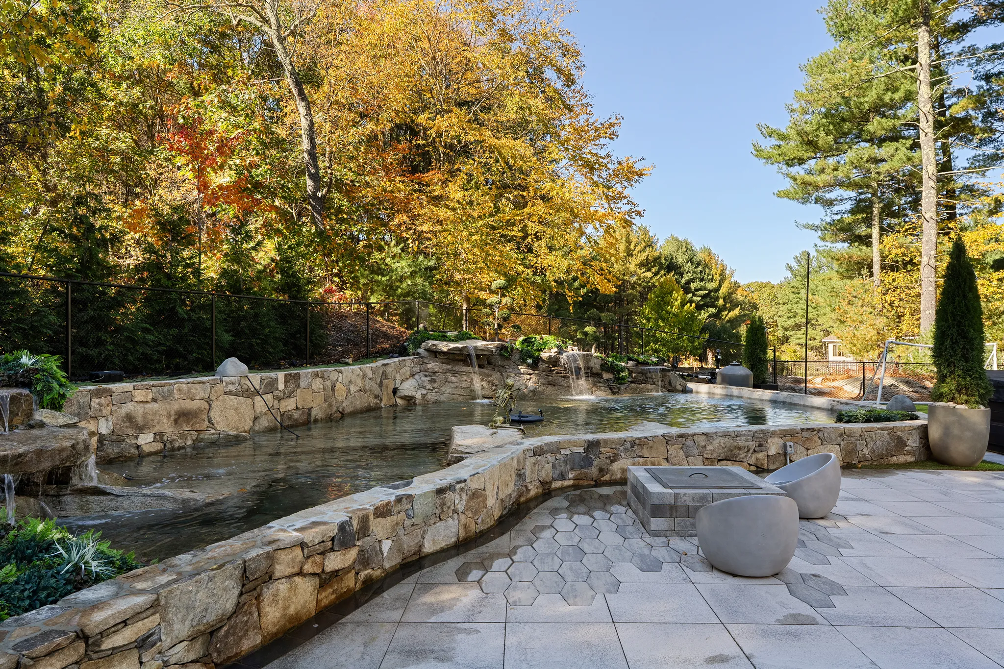 An outdoor living area features a stone-edged pond with waterfalls, a patio with a fire pit and seating, and fall foliage in the background.