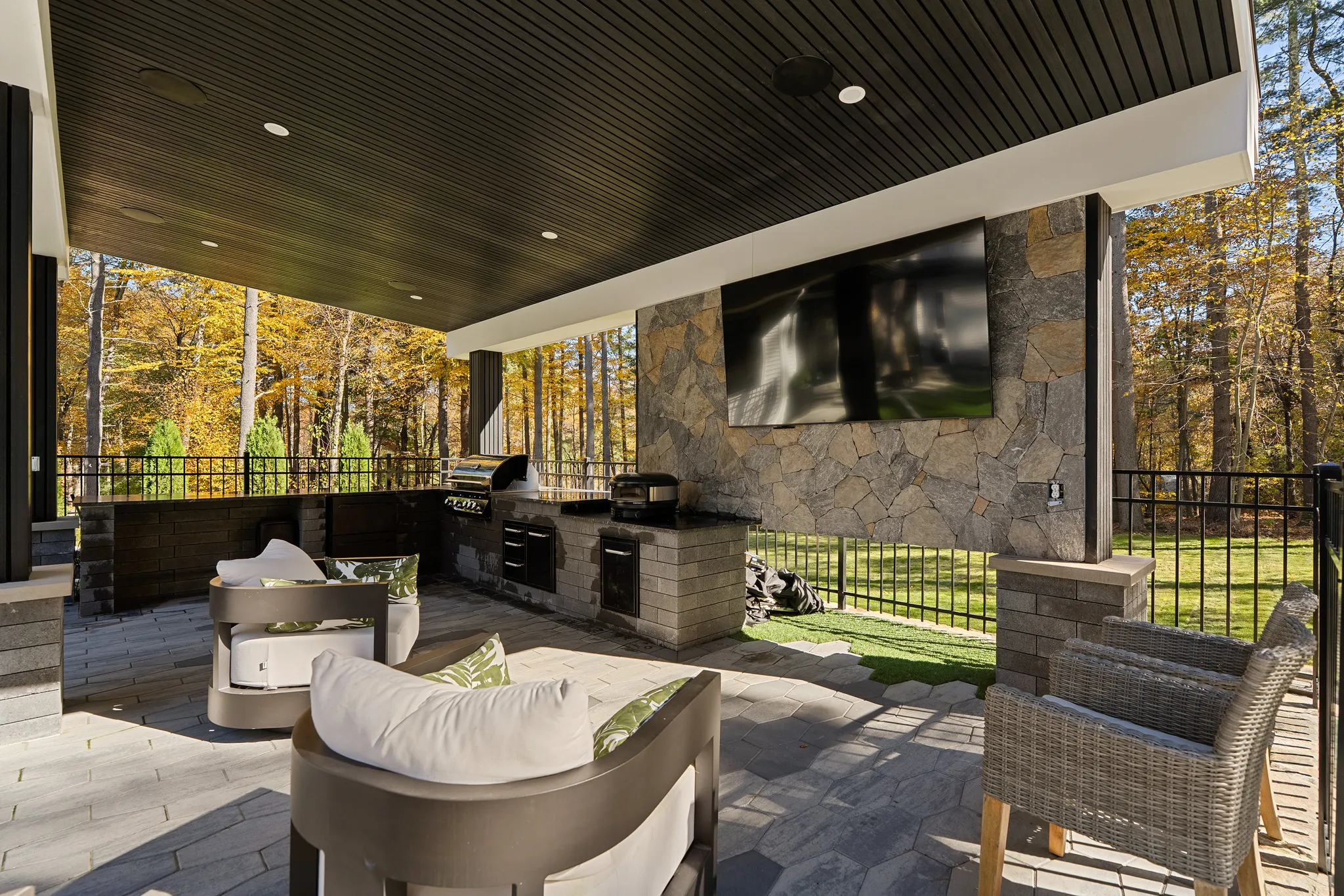 A covered outdoor patio features an outdoor kitchen with a grill, a mounted TV on a stone wall, and seating overlooking a fenced yard and autumn trees.