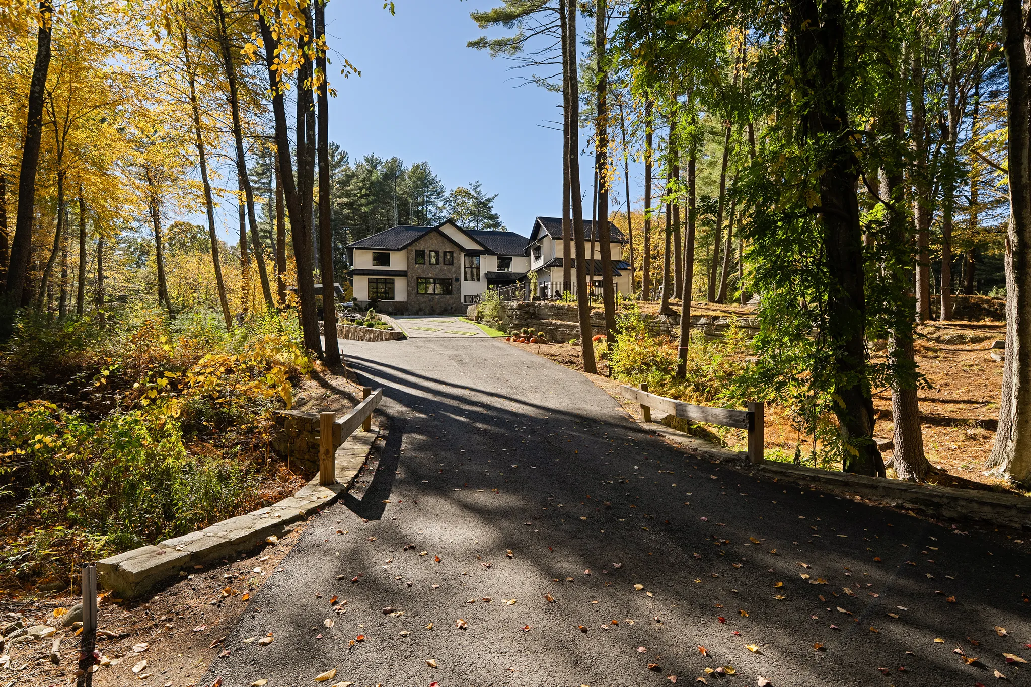 A modern house with a long paved driveway is situated in a wooded lot surrounded by autumn trees.