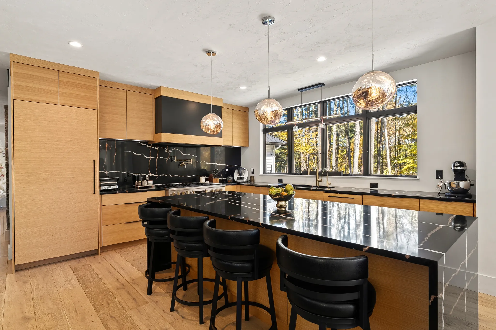 A modern kitchen features a black marble island with seating, wood cabinetry, and large windows overlooking trees.