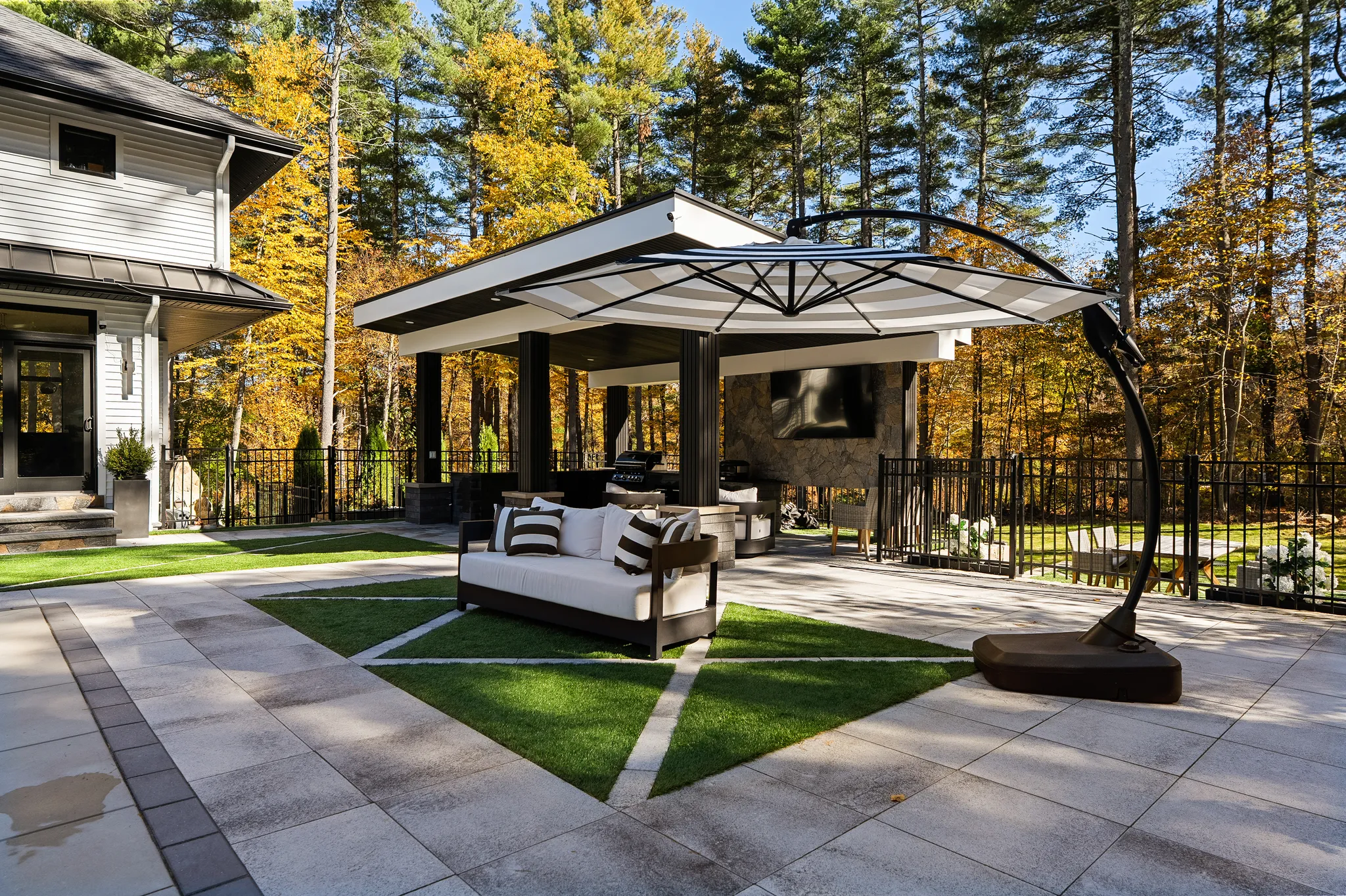 An exterior view of a modern patio featuring an outdoor kitchen, seating area, and dining space, surrounded by trees with autumn foliage.