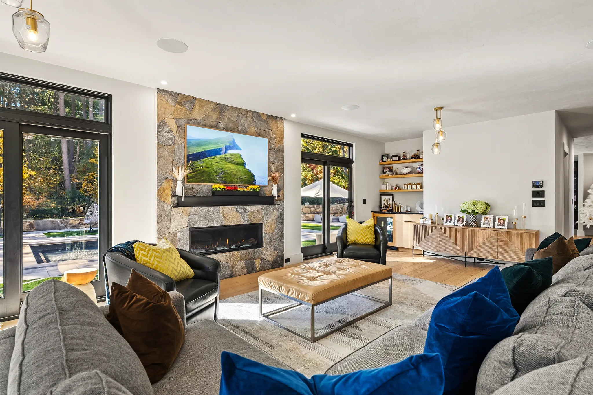 A modern living room with a stone fireplace, a large TV, a sectional sofa, and expansive windows looking out to a backyard with a pool.
