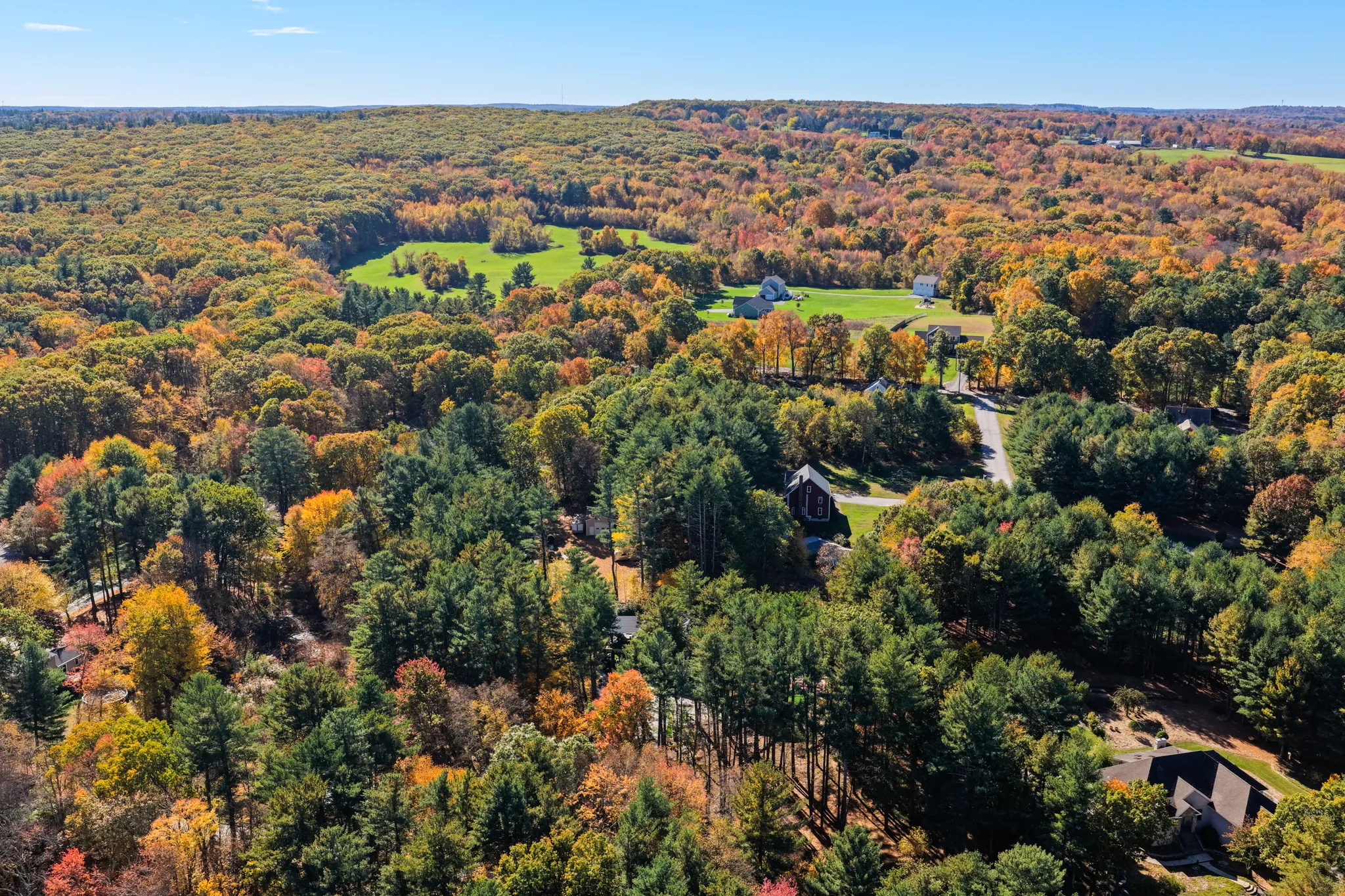 An aerial view displays a forested landscape with houses and open fields during the autumn season under a blue sky.