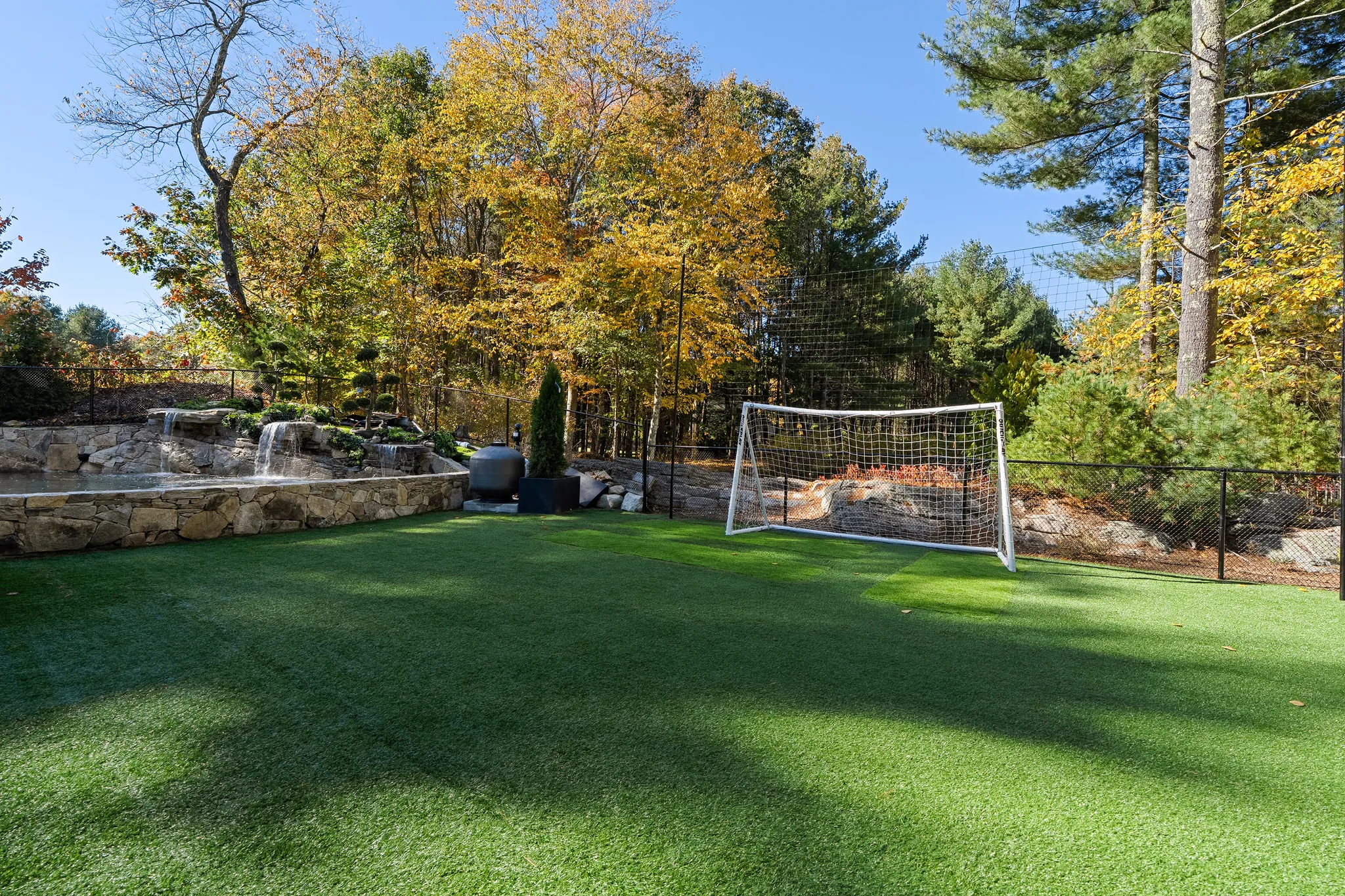 An exterior view of a backyard featuring an artificial turf soccer field, a waterfall feature, and trees with autumn foliage.