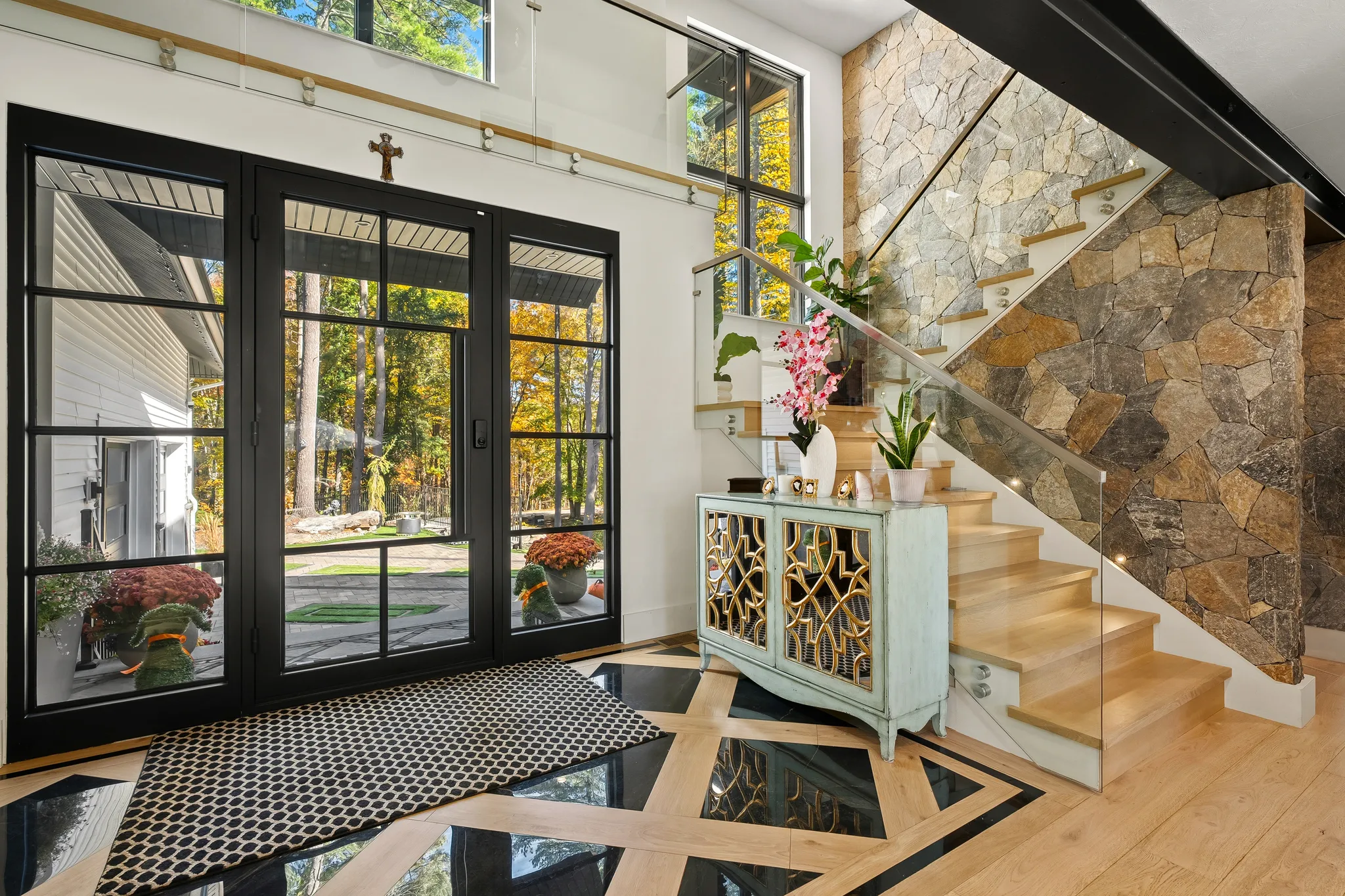 A modern entryway features a wooden staircase with glass railings, a stone accent wall, and large glass doors opening to an outdoor view.