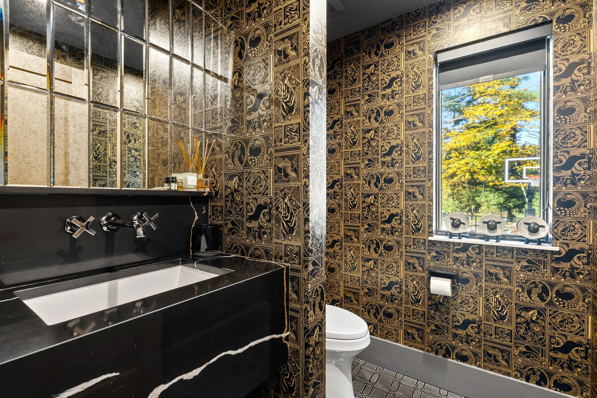 A powder room features black and gold patterned wallpaper, a black marble vanity with a wall-mounted faucet, a segmented mirror, and a toilet next to a window.