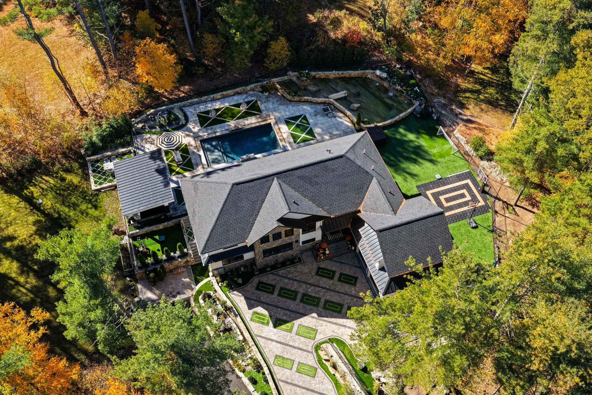 An aerial view of a large house with a swimming pool, sport court, and landscaped grounds surrounded by trees with autumn foliage.