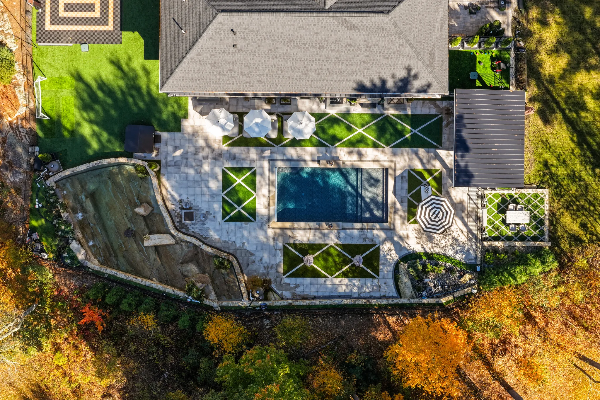 An aerial view of a house with a backyard featuring a swimming pool, patio, outdoor dining area, and a natural rock water feature, surrounded by colorful autumn trees.