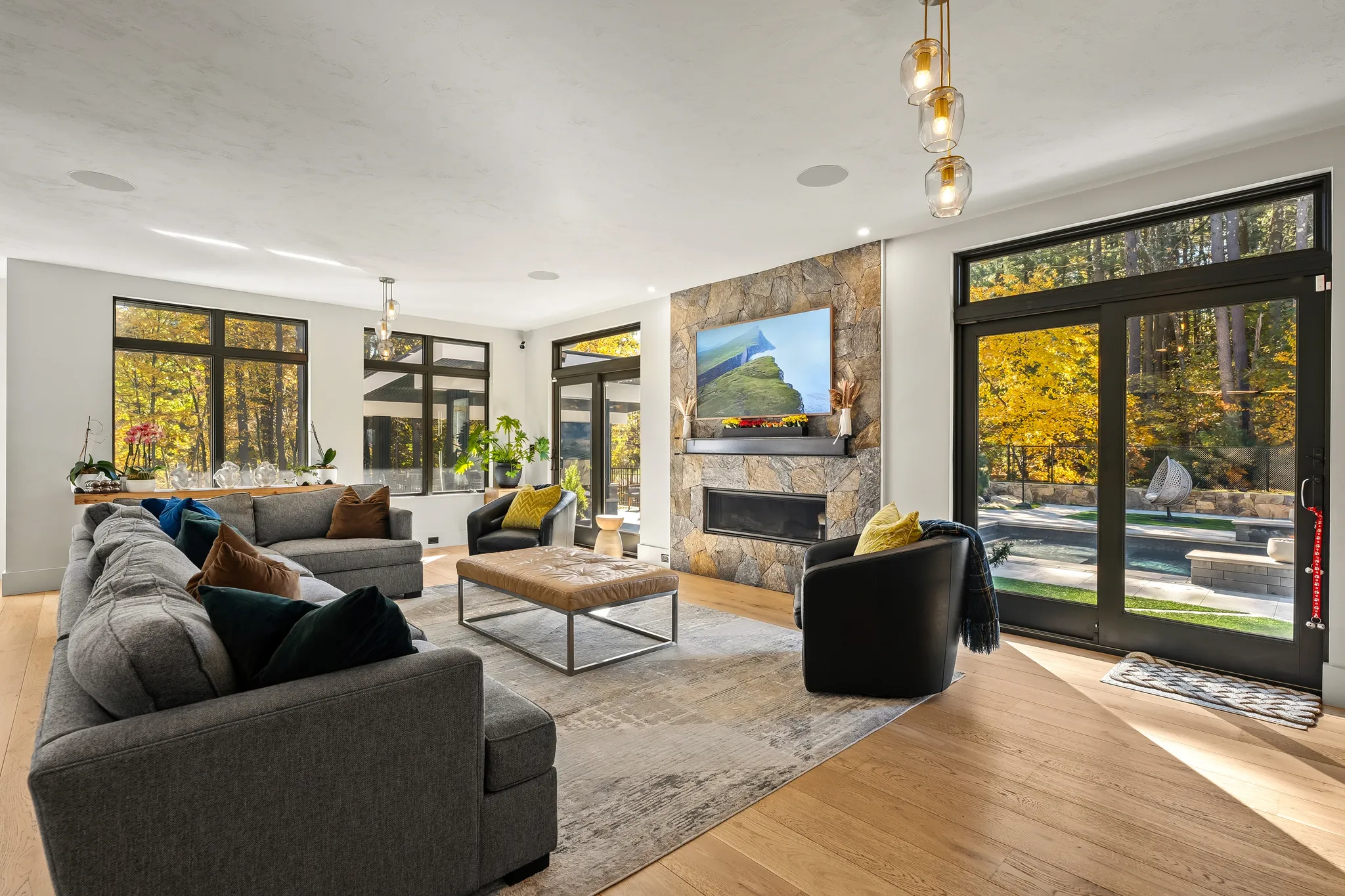 A modern living room features a stone fireplace, a large television, a sectional sofa, and expansive windows offering views of an autumn landscape.