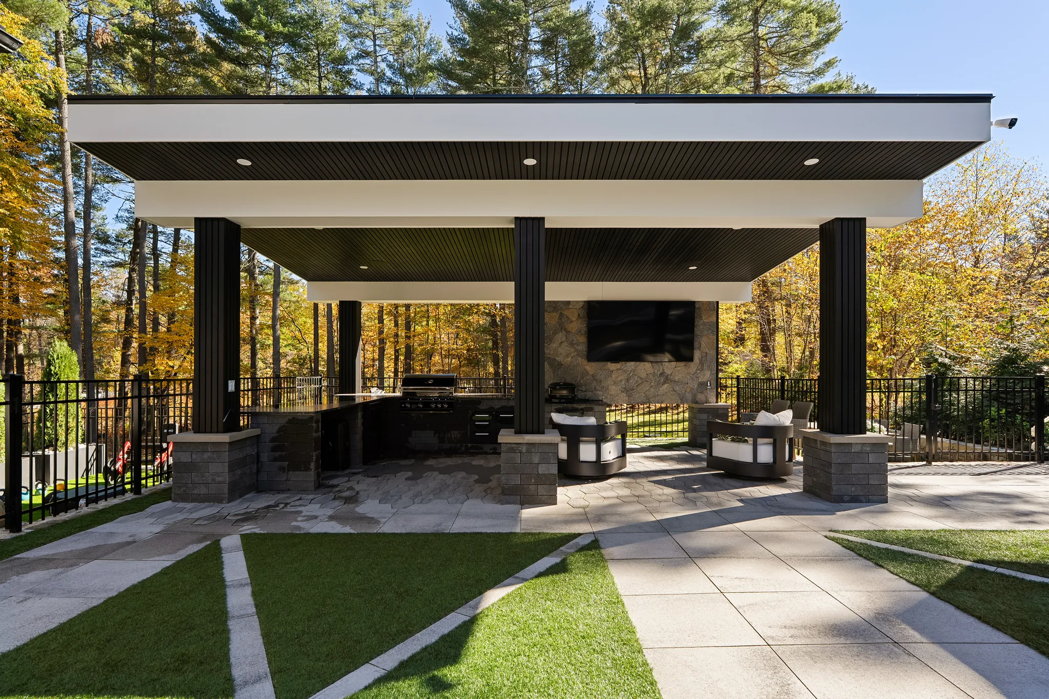 A modern outdoor living space features a covered patio with an outdoor kitchen, seating around a fire pit with a mounted television, and a yard with artificial turf, surrounded by trees with autumn foliage.