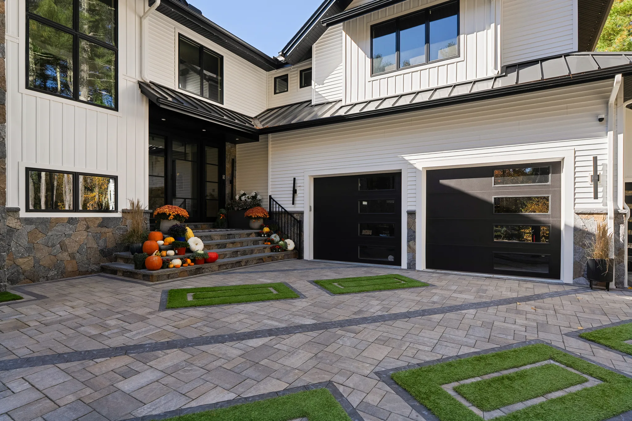 The front exterior of a modern white house with black garage doors and autumn decorations on the steps.