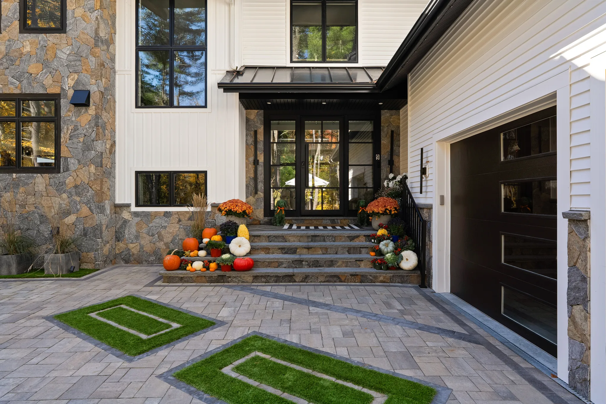 The front exterior of a modern house features stone cladding, white siding, a black front door, and a paver driveway with seasonal fall decorations.