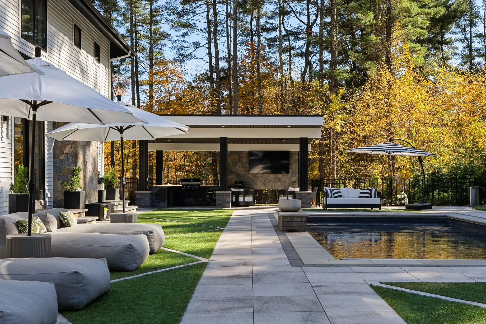 A spacious backyard features a swimming pool, an outdoor kitchen under a covered structure, a patio, and lounge chairs next to a modern house.