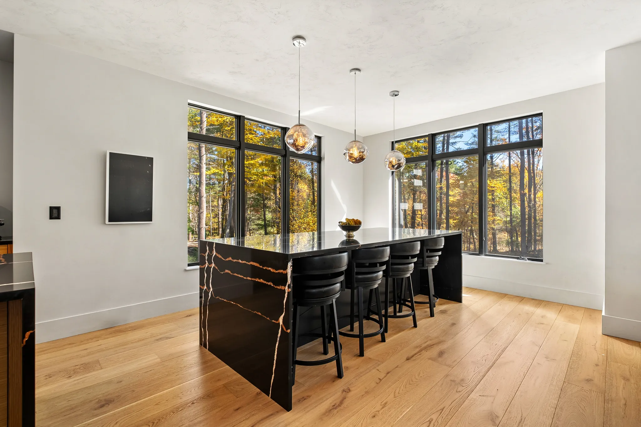 A modern kitchen features a large black island with bar stools, hardwood floors, and large windows overlooking a forest.