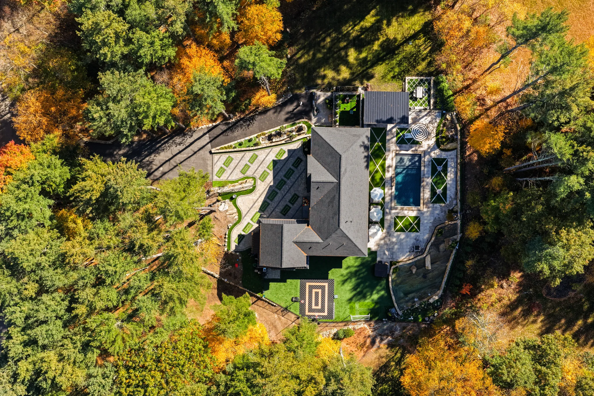 An aerial view of a house with a swimming pool, patio, and patterned driveway, surrounded by trees with autumn foliage.