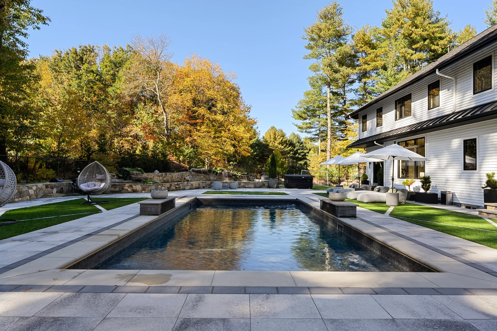 A backyard with a rectangular swimming pool, a patio area with seating, a hot tub, and a white house, surrounded by trees with autumn foliage under a clear sky.