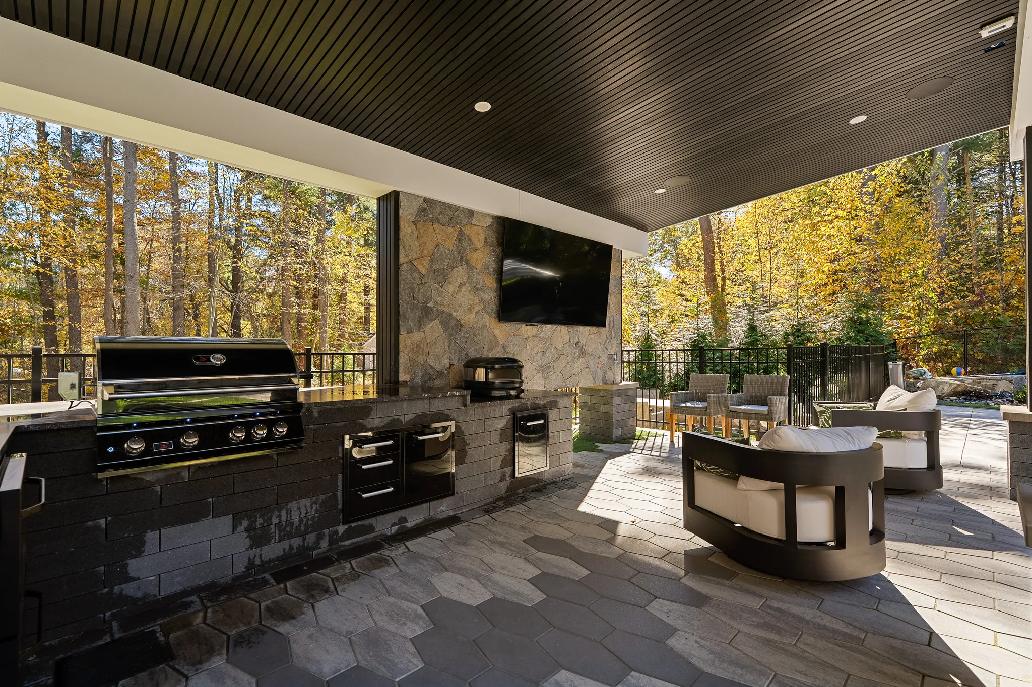 An outdoor patio features a fully equipped kitchen with a grill, a mounted television, and lounge seating, all overlooking a vibrant autumn forest.