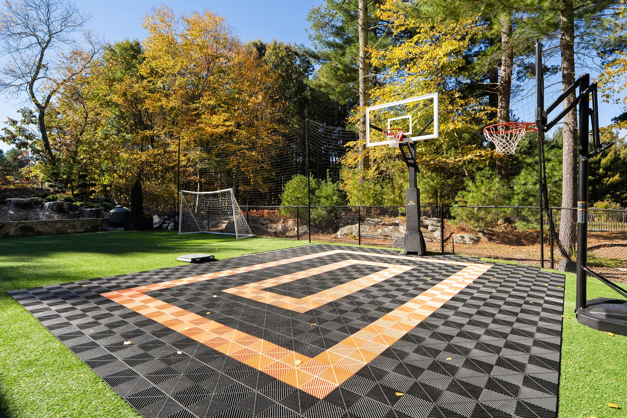 An outdoor sports court featuring two basketball hoops, a soccer goal, and a waterfall, surrounded by trees with autumn foliage.