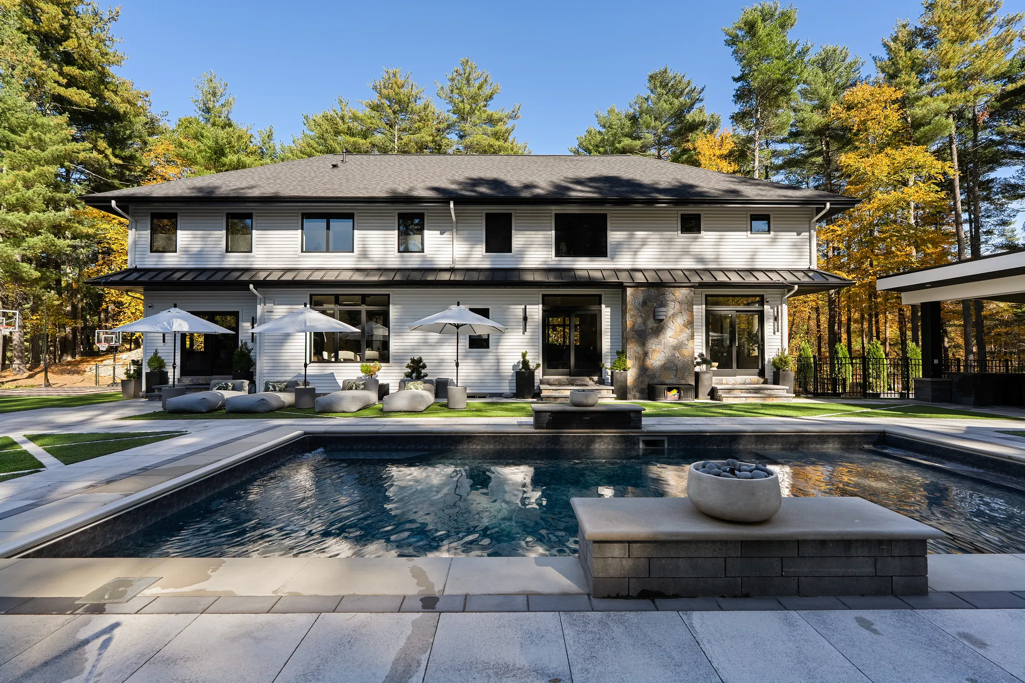 The back of a modern house features a swimming pool, a patio with outdoor seating, and a basketball court on the side.