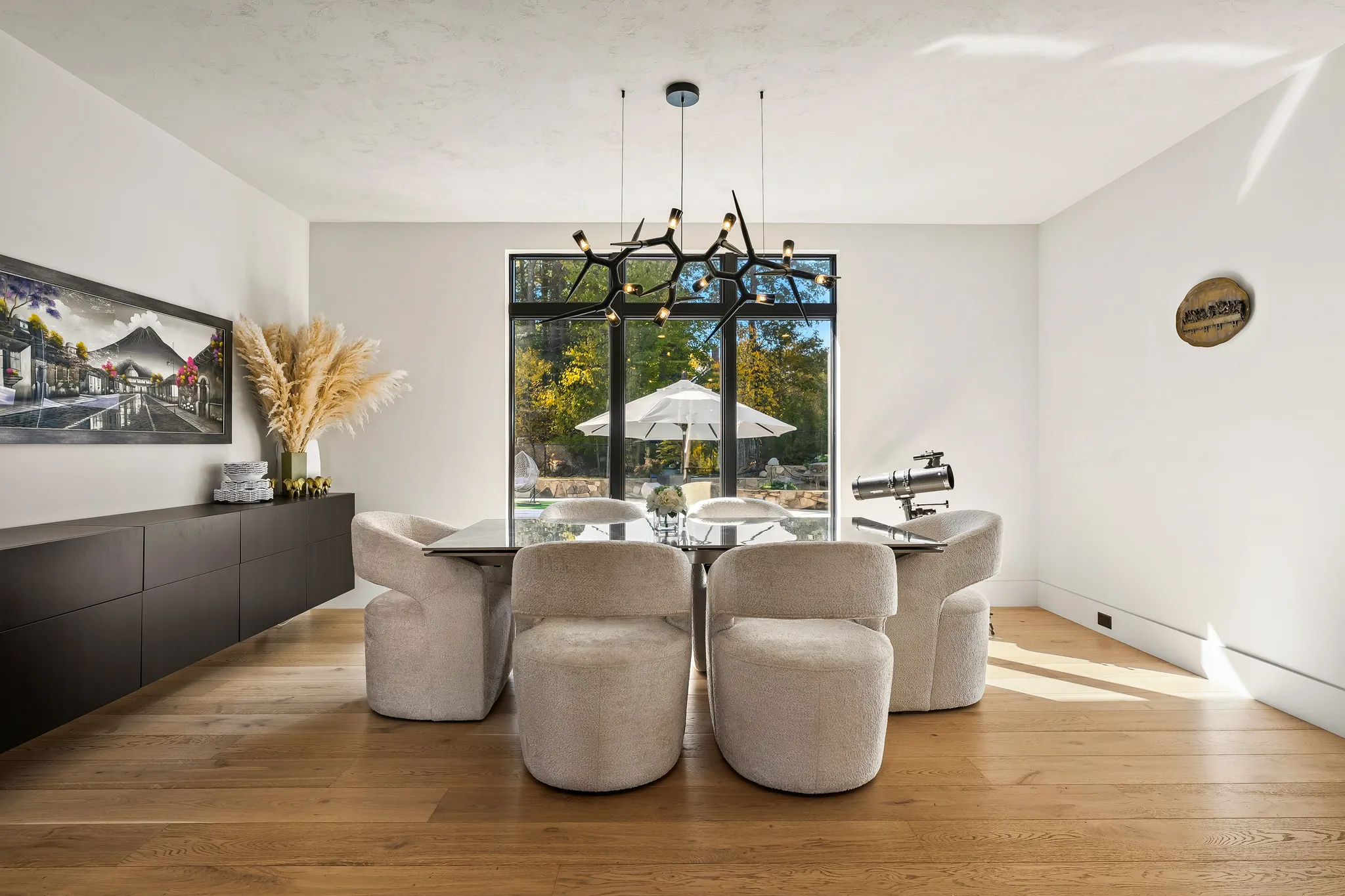A modern dining room features a long dining table with upholstered chairs, hardwood floors, a dark sideboard with art, and a large window looking out to a backyard.